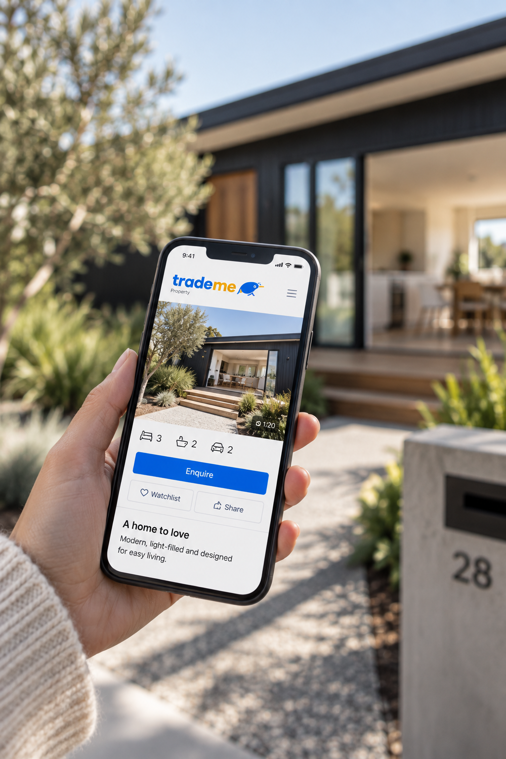 A person holding a smartphone with a real estate app displaying a modern house for sale, with a black exterior, front porch steps, garden, and open interior visible on the screen, outside a house.