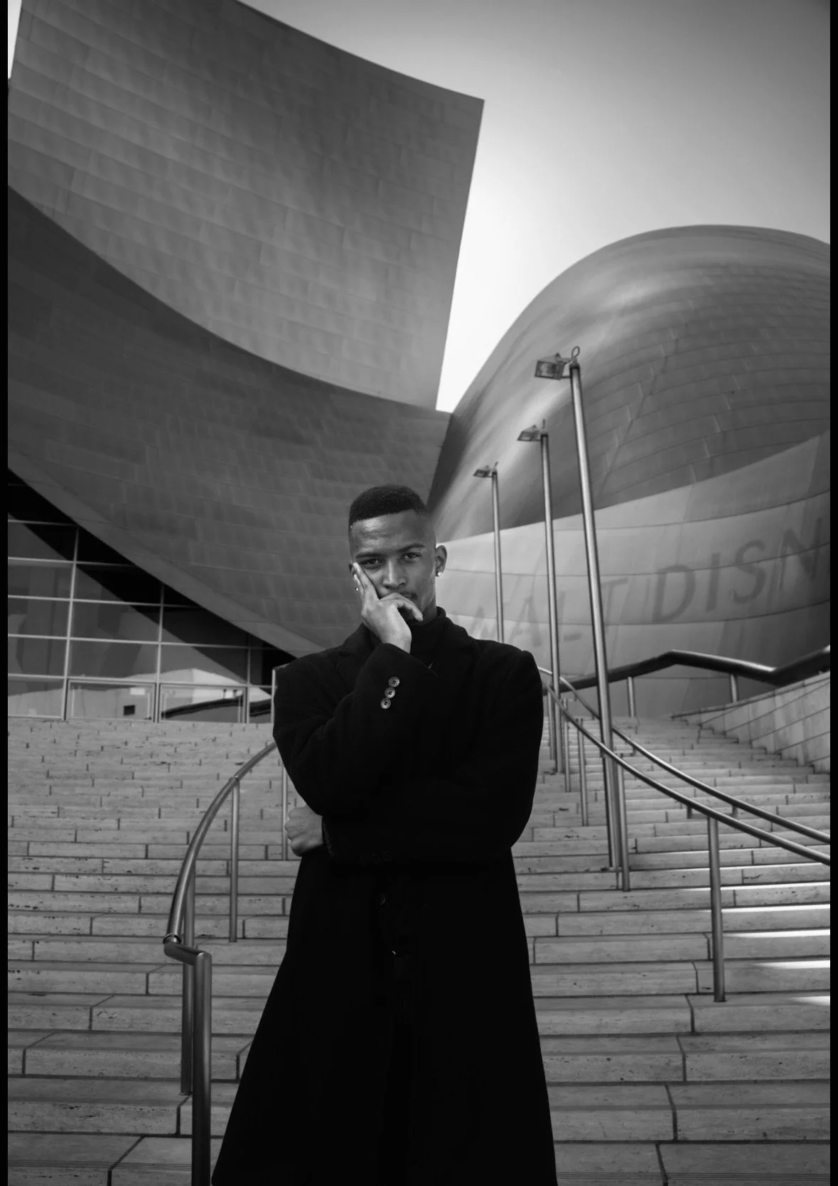 A man stands in front of a modern building with curved metallic architecture, steps, and railings, wearing a dark coat and resting his hand on his face.