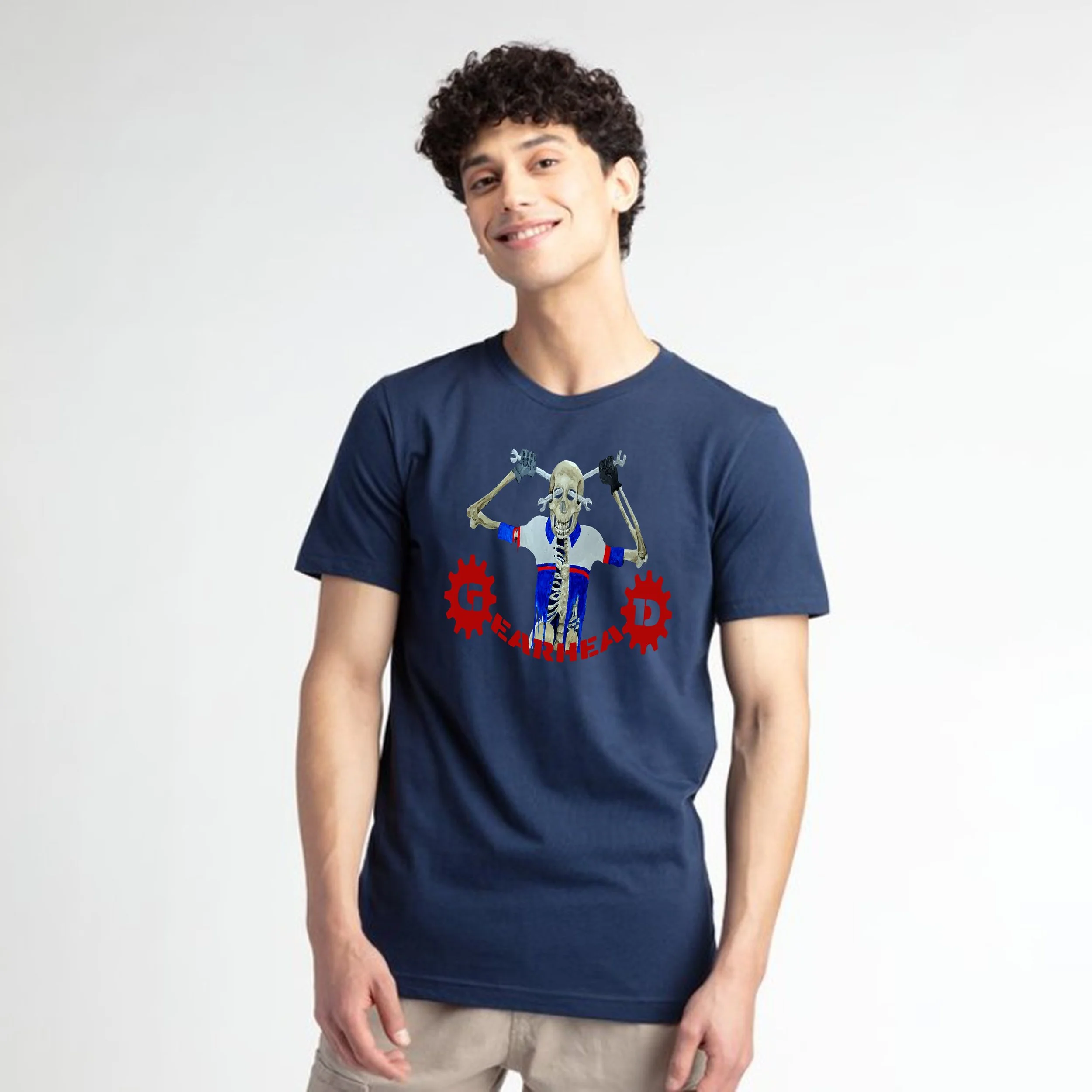 A young man with curly dark hair and light skin, smiling, wearing a navy blue T-shirt featuring a skeleton and gear graphic designs, and beige pants, standing against a plain white background.