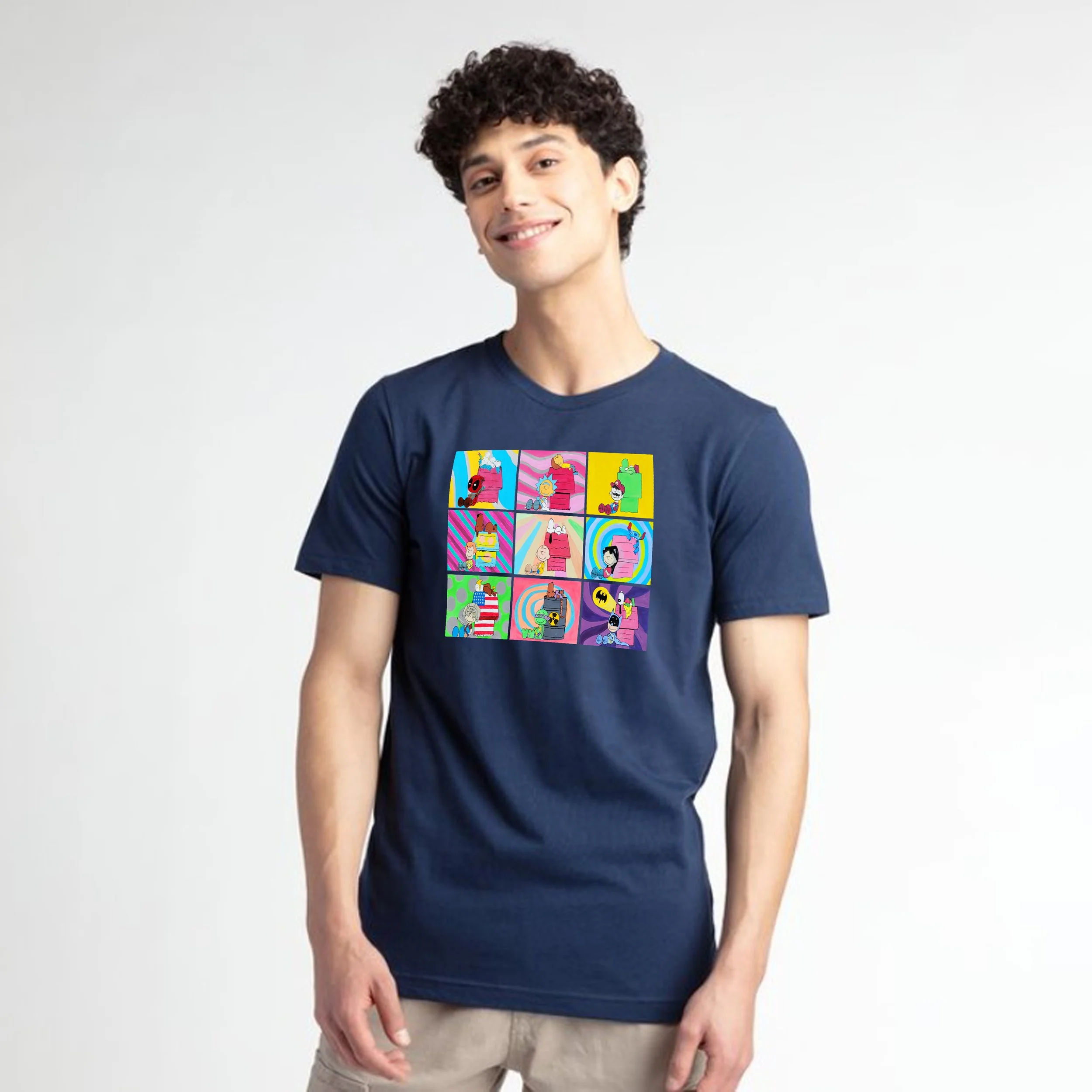 Young man with curly hair smiling, wearing a navy blue graphic t-shirt and light-colored pants, standing against a plain light gray background.