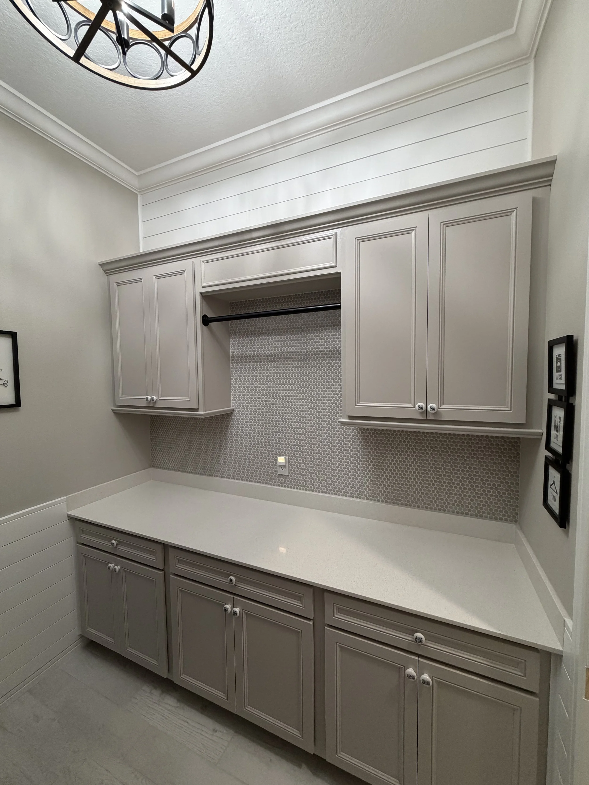 Kitchen cabinetry with cream-colored upper and lower cabinets, a white countertop, and a textured tile backsplash. A clock is mounted on the ceiling in the upper left corner.