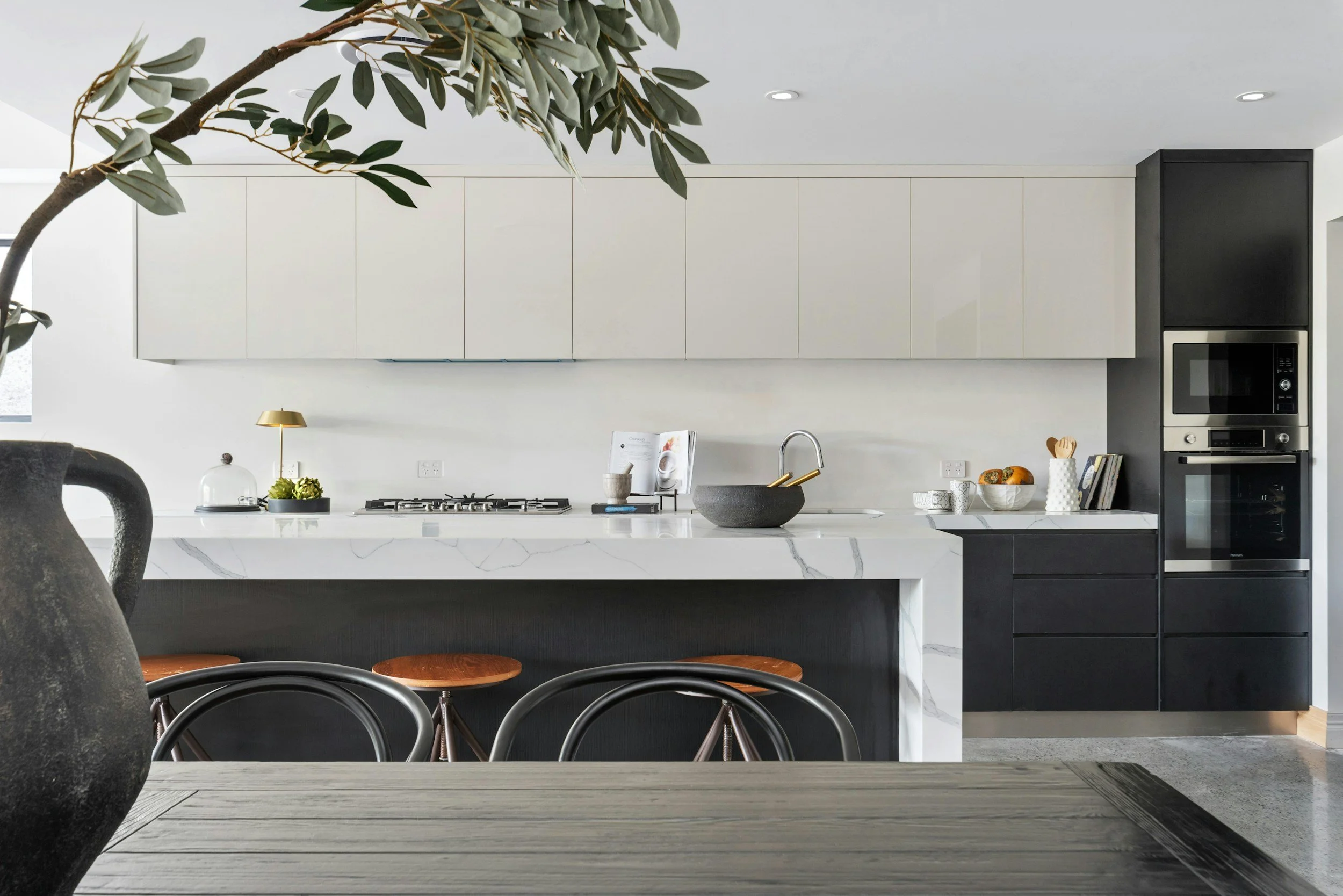 Modern kitchen with white and black cabinetry, marble countertop, and kitchen appliances. There is a leafy plant in the foreground and dining chairs in the foreground.
