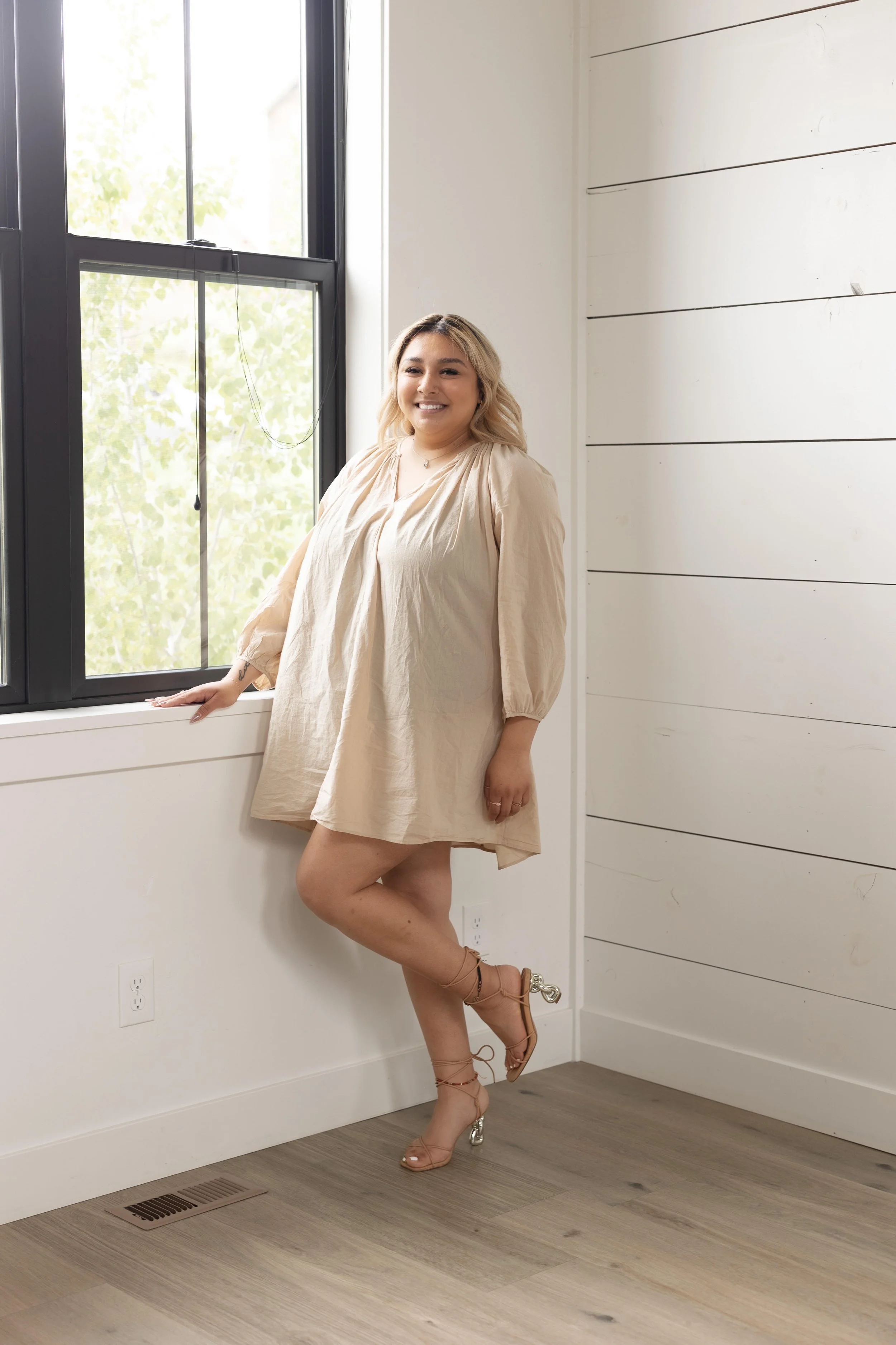 A woman with blonde hair smiling, wearing a beige dress and clear high-heeled shoes, standing next to a window with black framing in a bright room with white walls and wooden flooring.