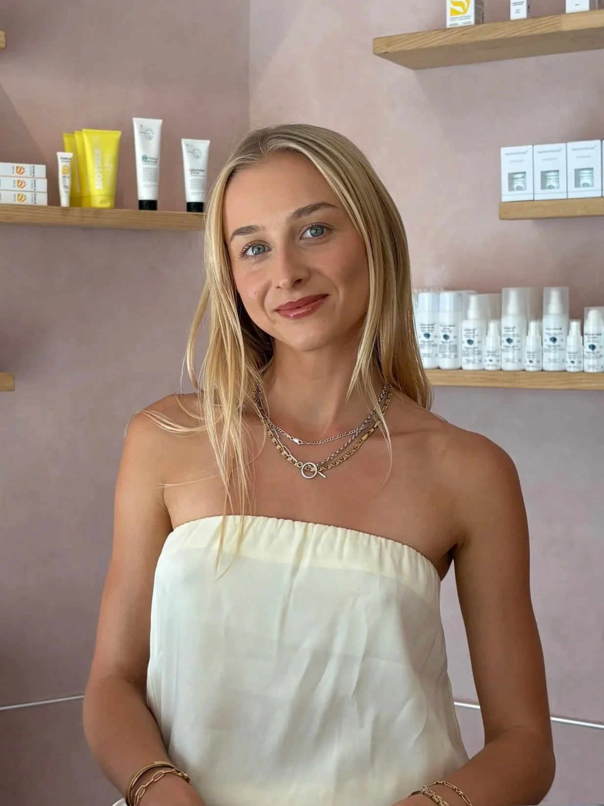 A young woman with long blonde hair, wearing a strapless cream-colored top and layered necklaces, stands in front of wooden shelves filled with skincare or beauty products in a retail or spa setting.