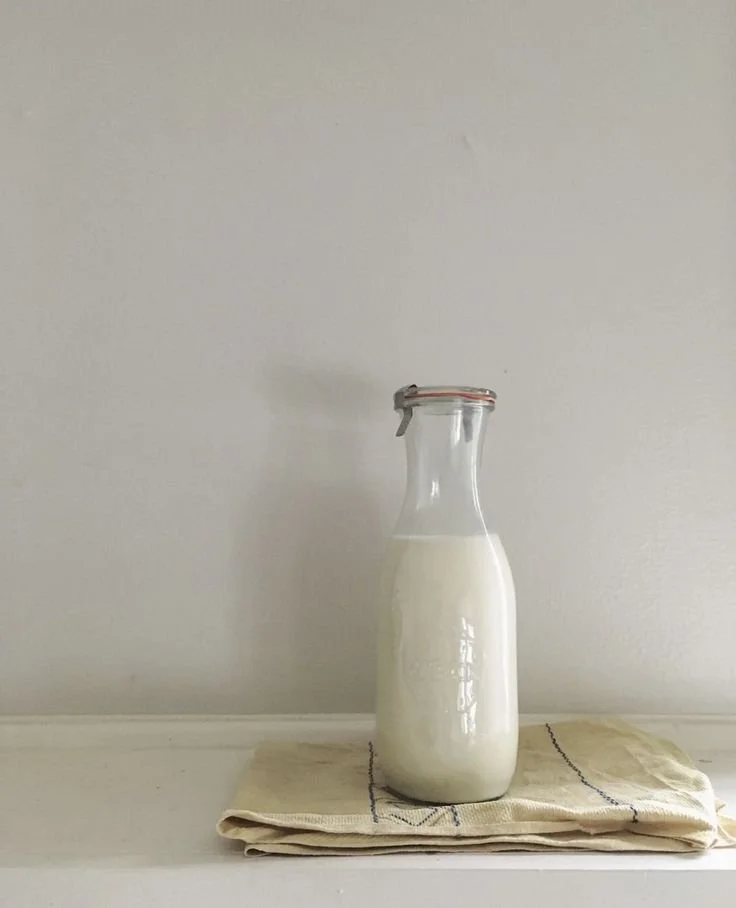 Glass bottle filled with milk placed on a beige cloth on a white surface against a plain wall.