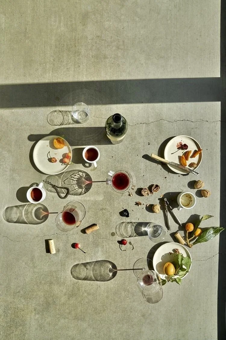 Top-down view of a table with glasses of red wine, bottles, plates with fruit and snacks, corks, and decorative items, all casting shadows on a concrete surface illuminated by sunlight.