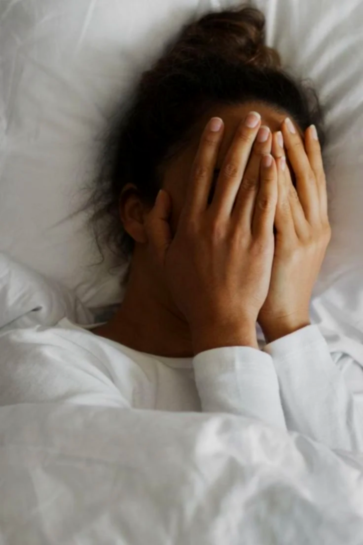 A woman lying in bed with her hands covering her face, possibly experiencing stress or pain.