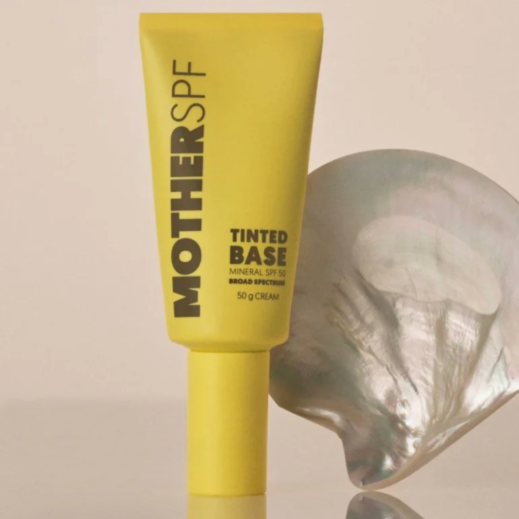 A yellow tube of Moisture SPF tinted base cream with broad spectrum SPF 50, standing upright on a flat surface next to a metallic seashell.