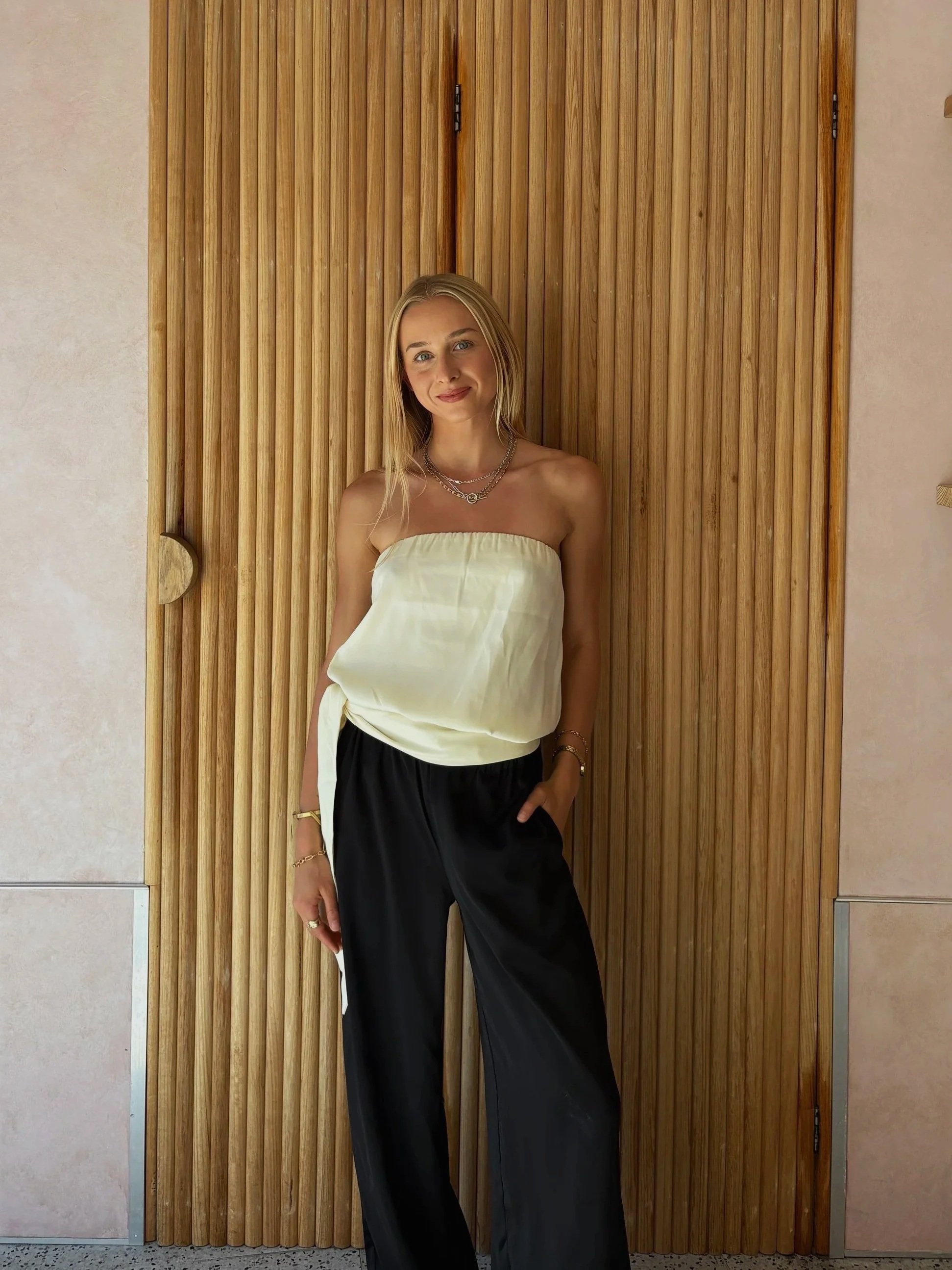 A young woman standing in front of a wooden door with vertical panels, wearing a strapless cream-colored top and black pants, with jewelry including necklaces, bracelets, and a ring.