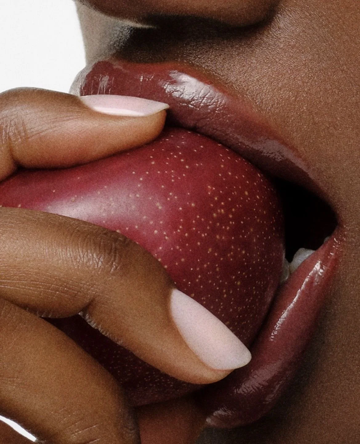 Close-up of a person with dark skin licking a red strawberry with yellow seeds, with lips slightly parted and tongue visible.