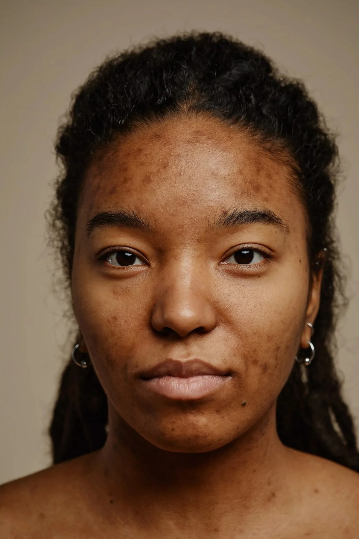 Close-up portrait of a young woman with dark skin and natural curly hair, wearing earrings, and looking directly at the camera against a plain background.