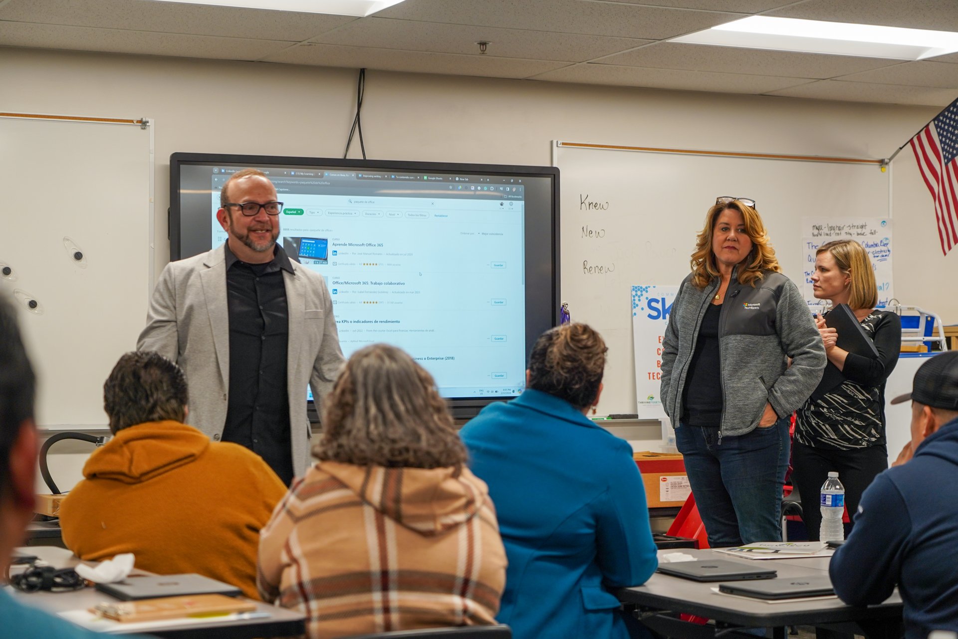 A classroom or workshop setting with three women and several seated participants. One woman stands near a digital screen displaying a web page, smiling, while the other two women stand nearby, one with arms crossed and the other holding a tablet. The room has a whiteboard, an American flag, and supplies like water bottles, laptops, and notebooks.