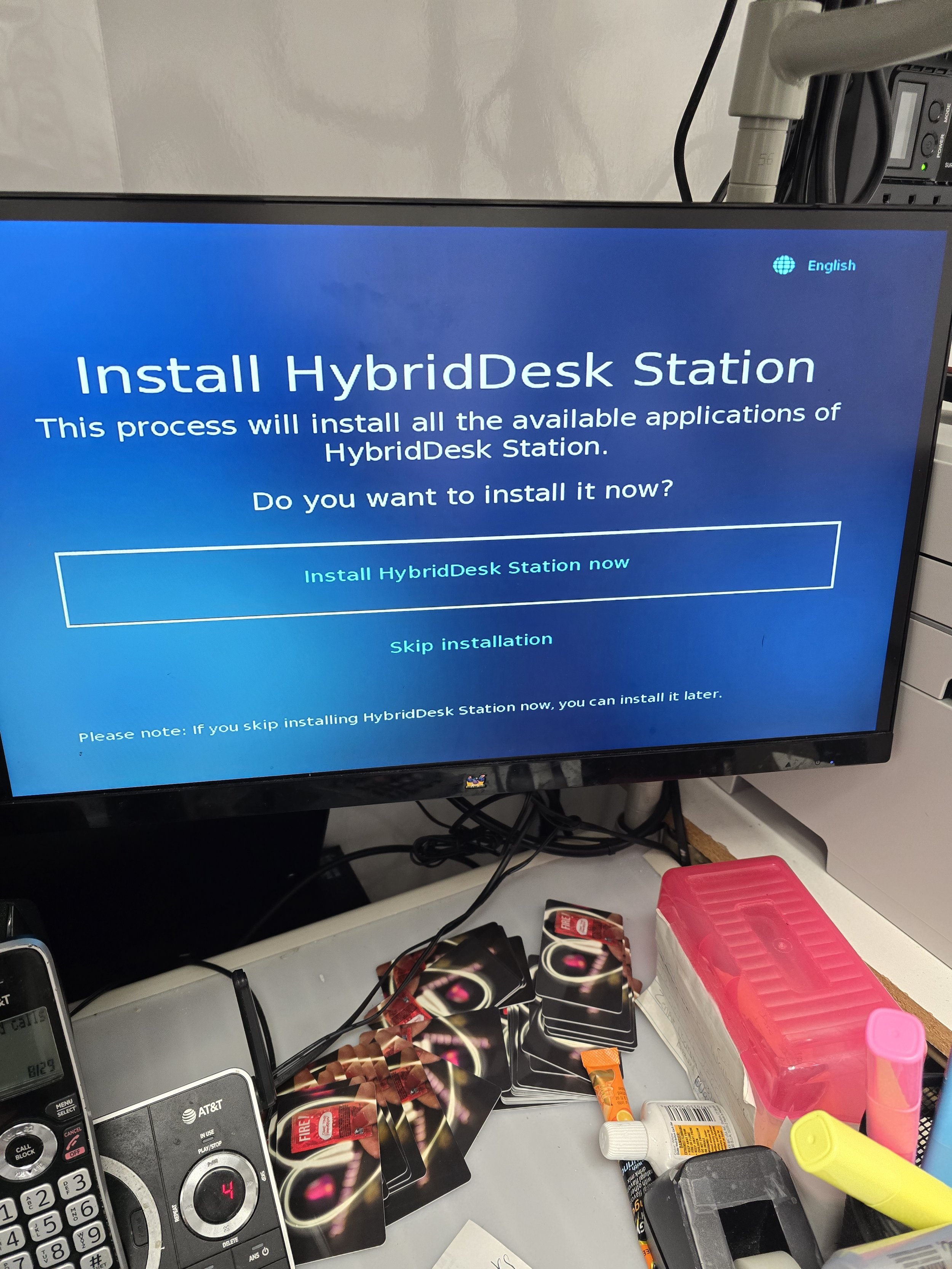 A computer monitor displaying an installation screen of HybridDesk Station with options to install or skip, surrounded by scattered cards, a pink container, a telephone, and various office supplies on a cluttered desk.