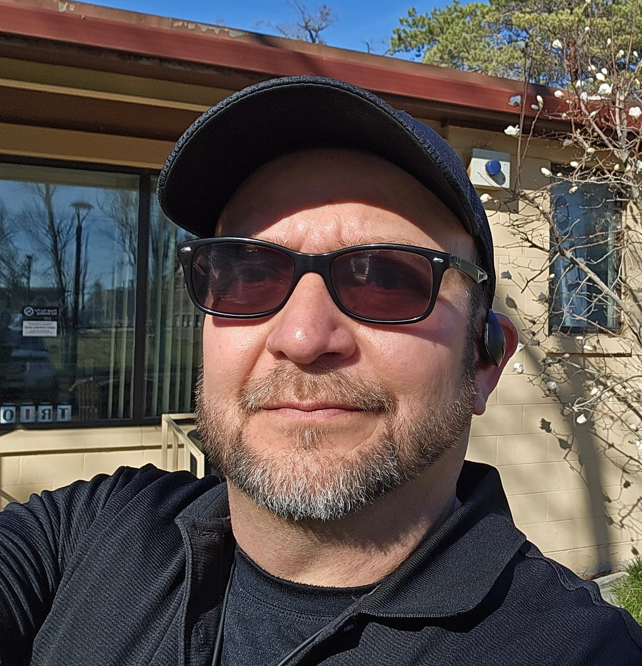 A man wearing sunglasses, a black cap, and a black shirt, smiling outdoors in front of a building with a large window and trees in the background.