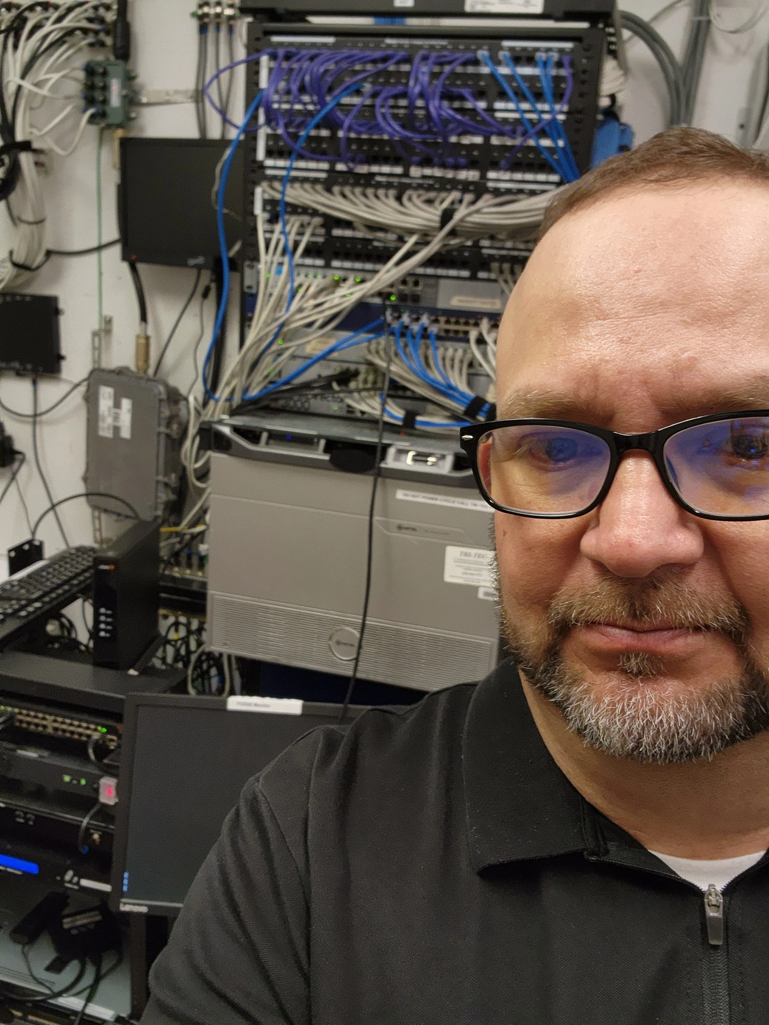 A man with glasses taking a selfie in a server room filled with network equipment, switches, and cables.