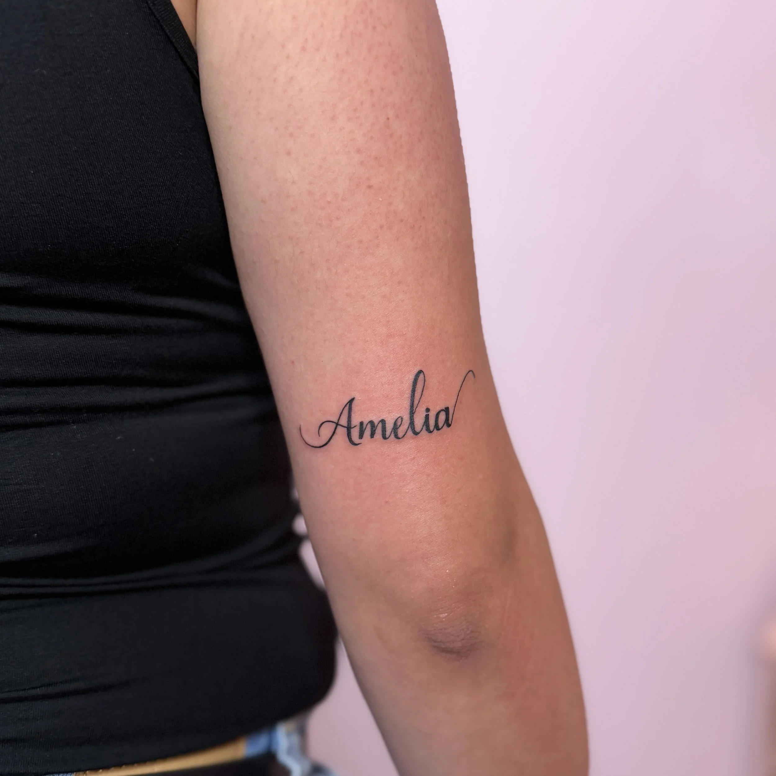 Close-up of a person's upper arm with a tattoo reading 'Amelia' in cursive, against a pink background.