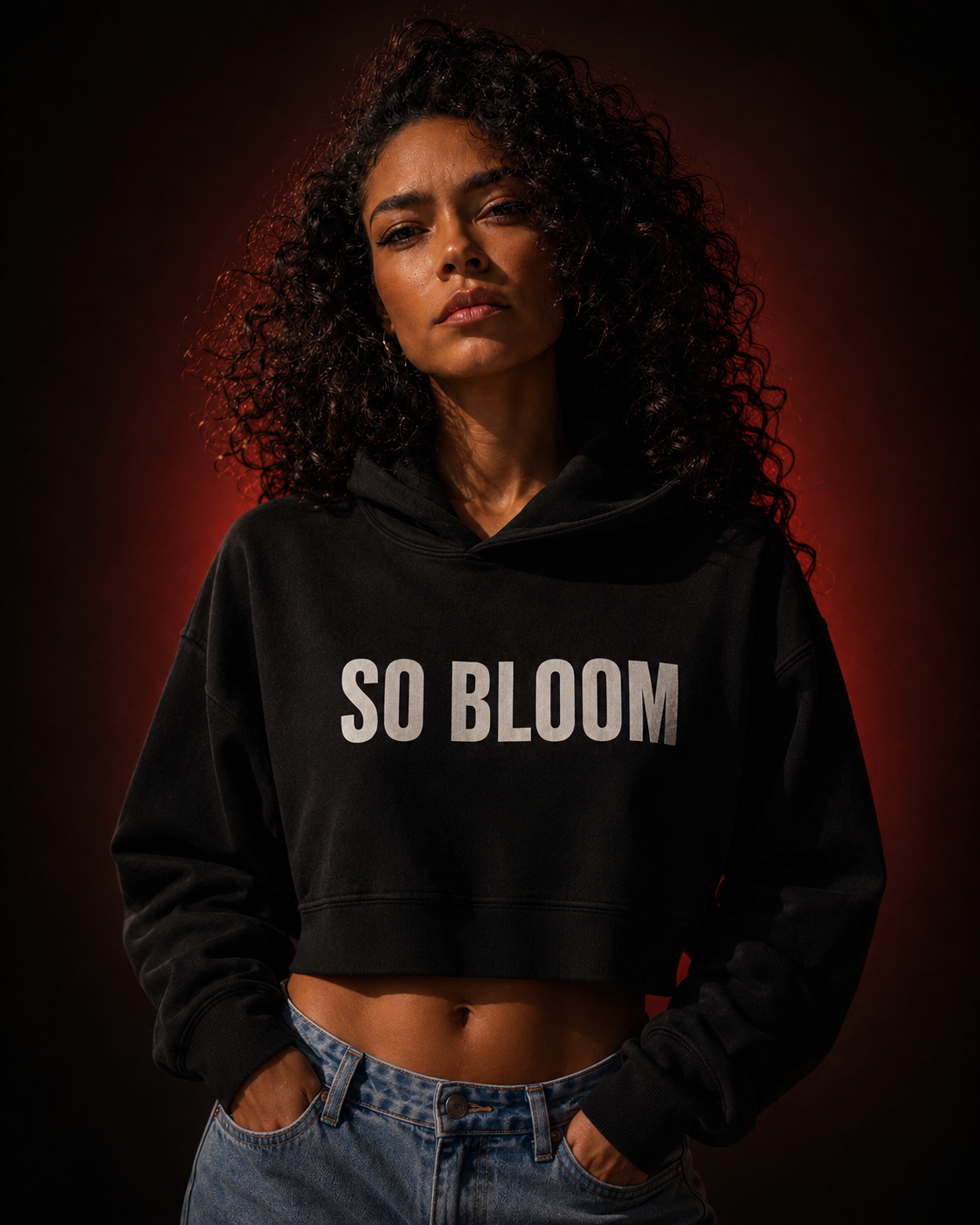 A young woman with curly hair wearing a black hoodie with white text that says 'SO BLOOM' and blue jeans, standing against a dark background with a red gradient.