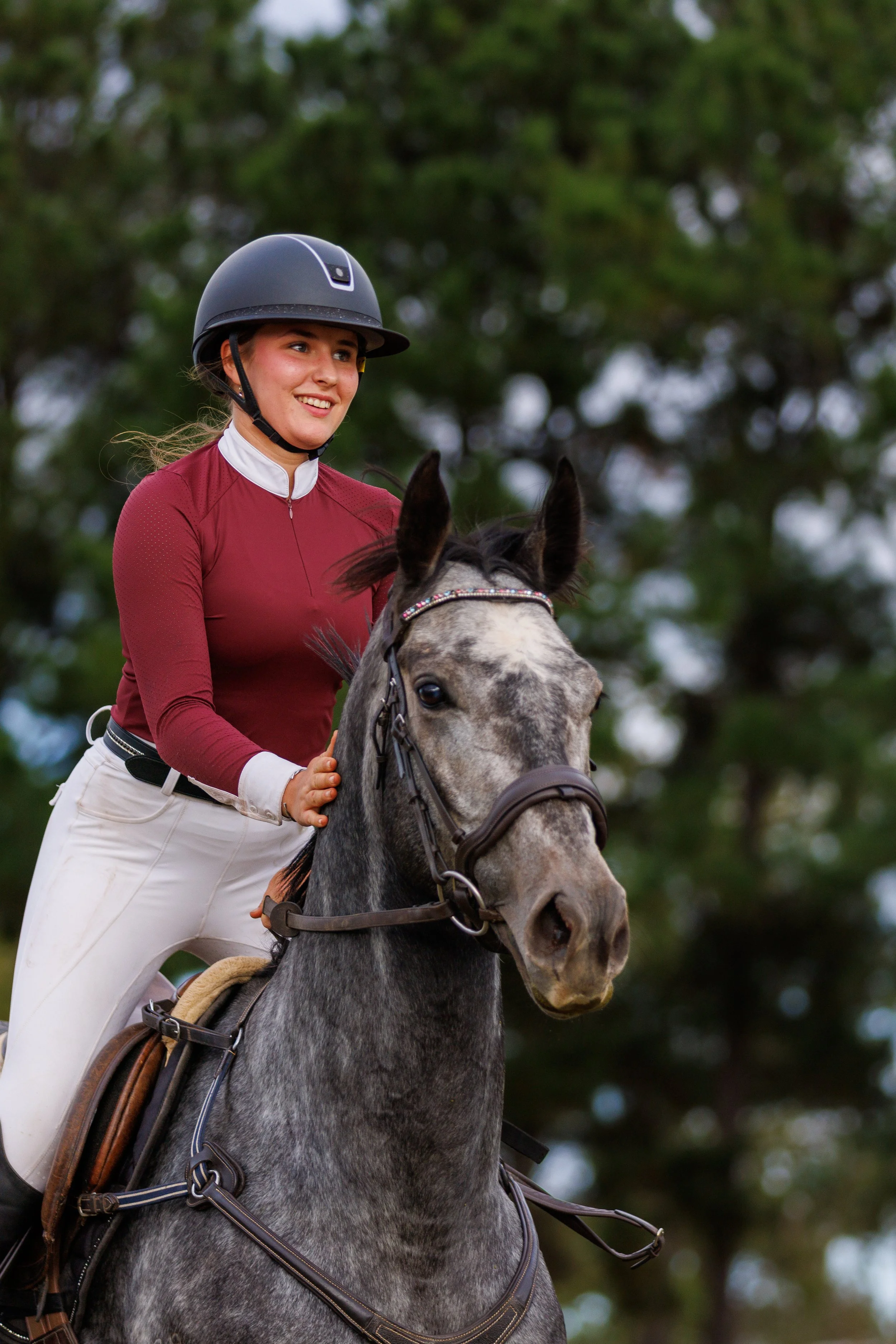 Young woman riding a gray horse outdoors, wearing a black riding helmet, maroon long-sleeve shirt, and white riding pants, smiling with trees in the background.