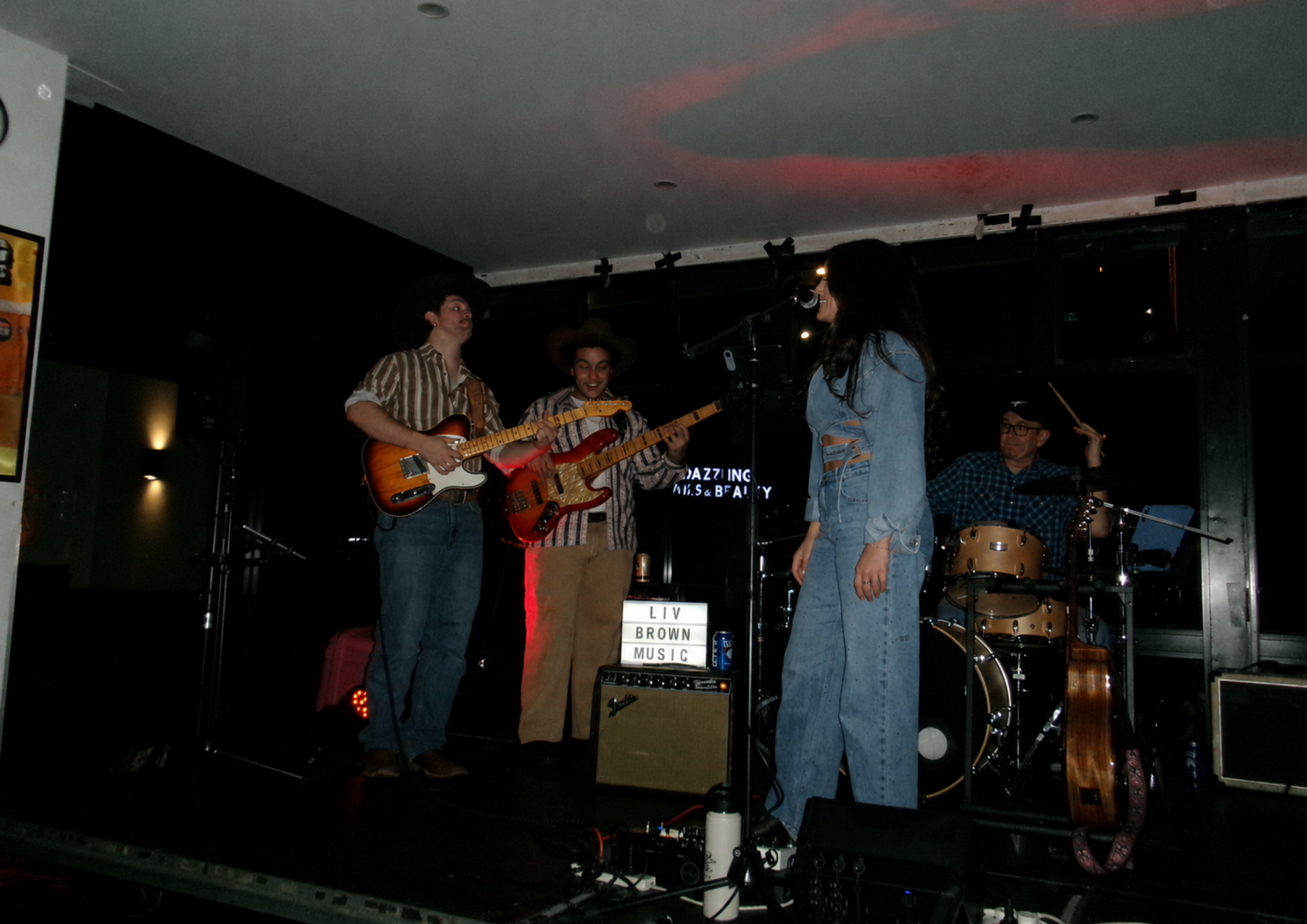 A group of four musicians performing on stage. Two of them are playing guitars, one is singing into a microphone, and another is playing drums in the background. The stage has a sign that reads 'Liv Brown Music'.