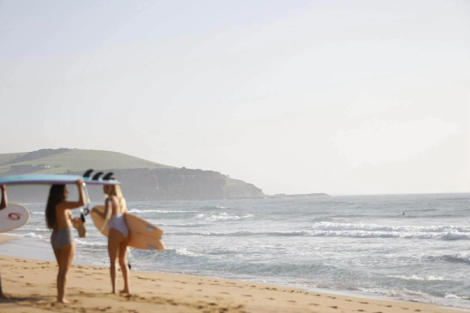 Two women walking on the beach with surfboards, ocean waves in the background, and a green hillside in the distance.