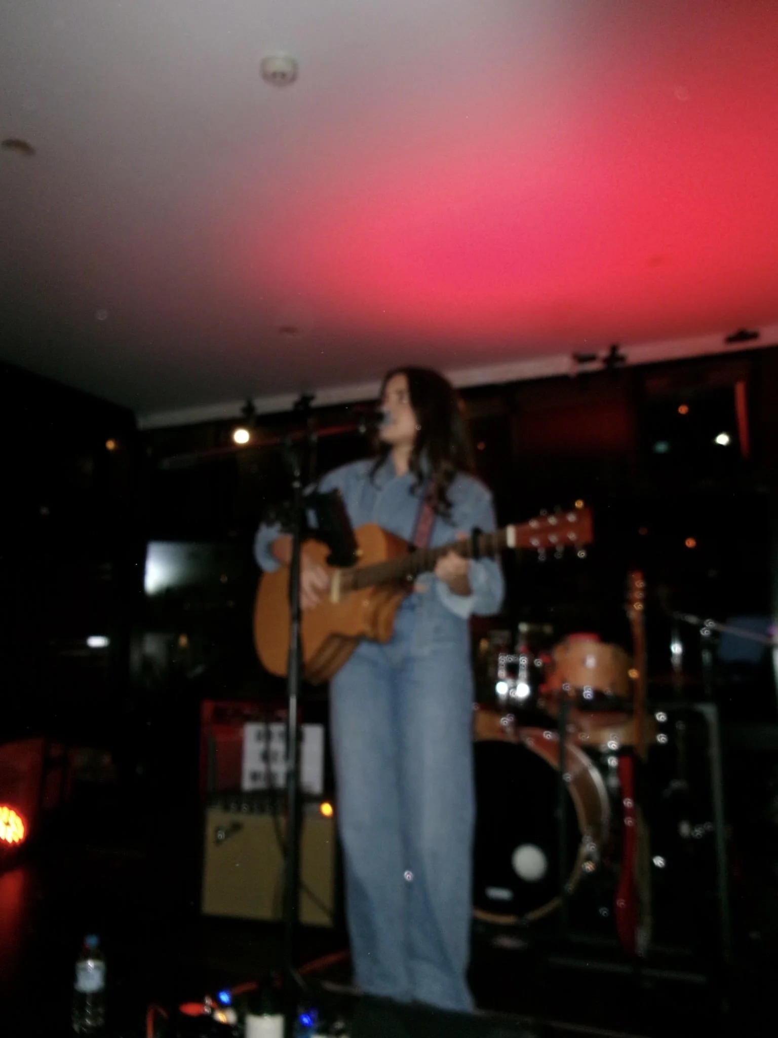 A woman playing guitar and singing on stage inside a dimly lit venue with a drum set in the background and a prominent red light on the ceiling.