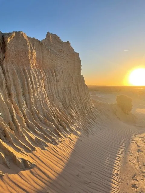 Sand dunes with ridges cast in shadow, near a cliff, at sunset with a clear sky.