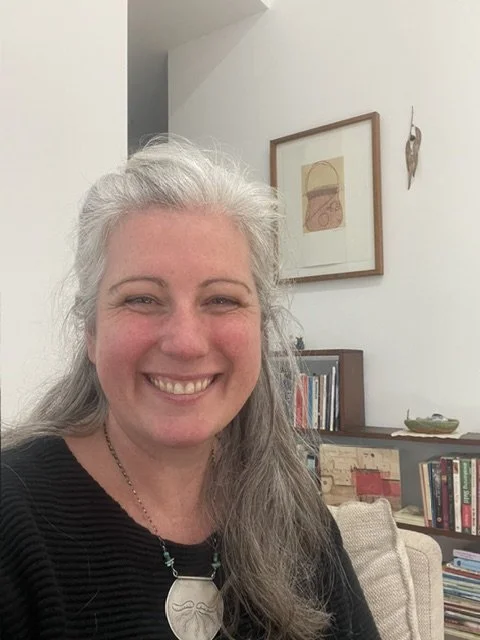 A woman (Sarah) smiling with gray hair in a black top sitting in a cozy room with a bookshelf and framed artwork on the wall behind her.