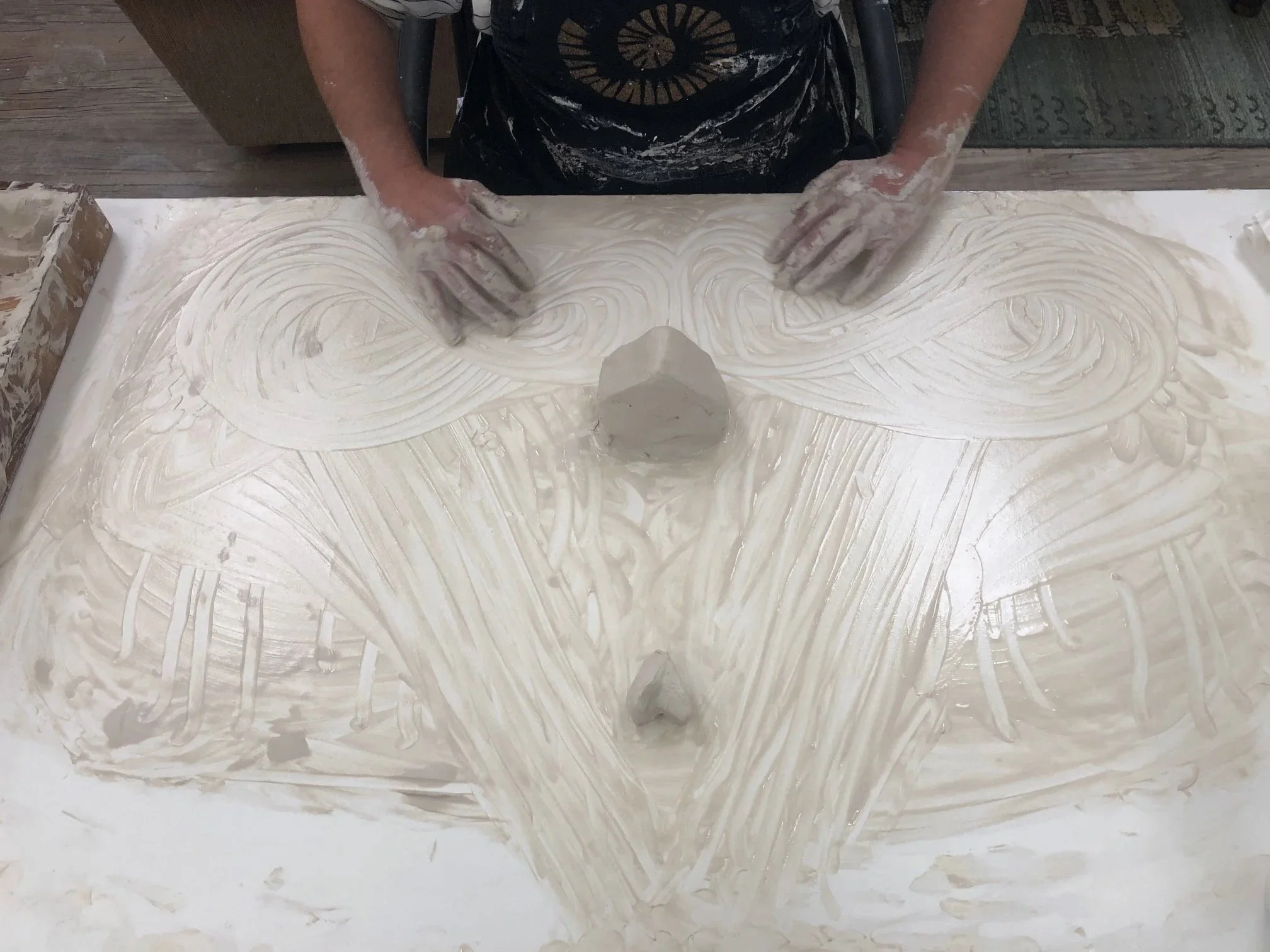 Person with art apron leaning over table covered in clay. Swirling design.