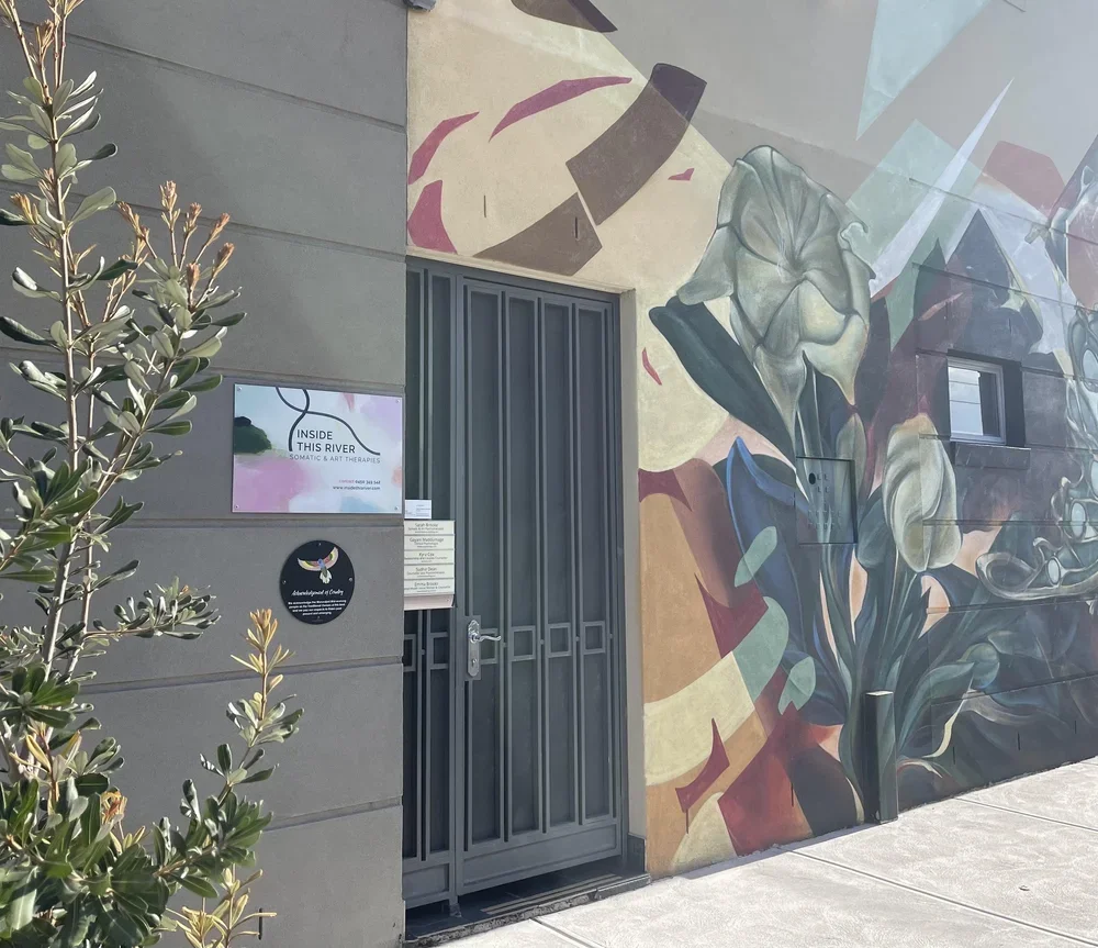 Street view of a building with a colorful mural featuring a woman with flowers and abstract shapes, a gray metal door, a small window, and a sign for Inside This River: Somatic & Art Therapies. A plant is in the foreground.