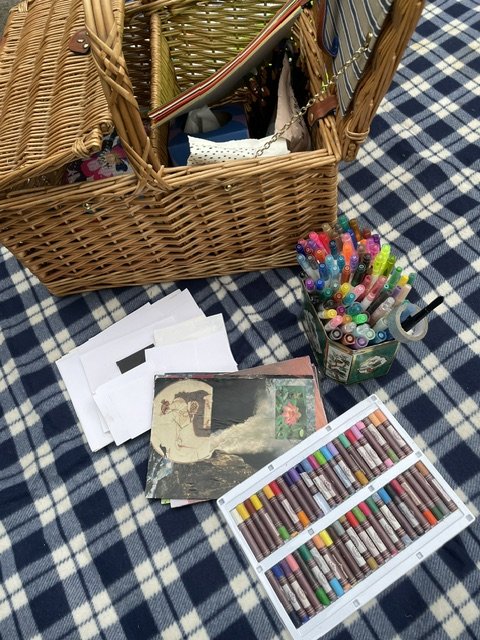 A picnic blanket with a wicker basket containing art therapy supplies, collage materials and craft paper. Outdoor sunlight.