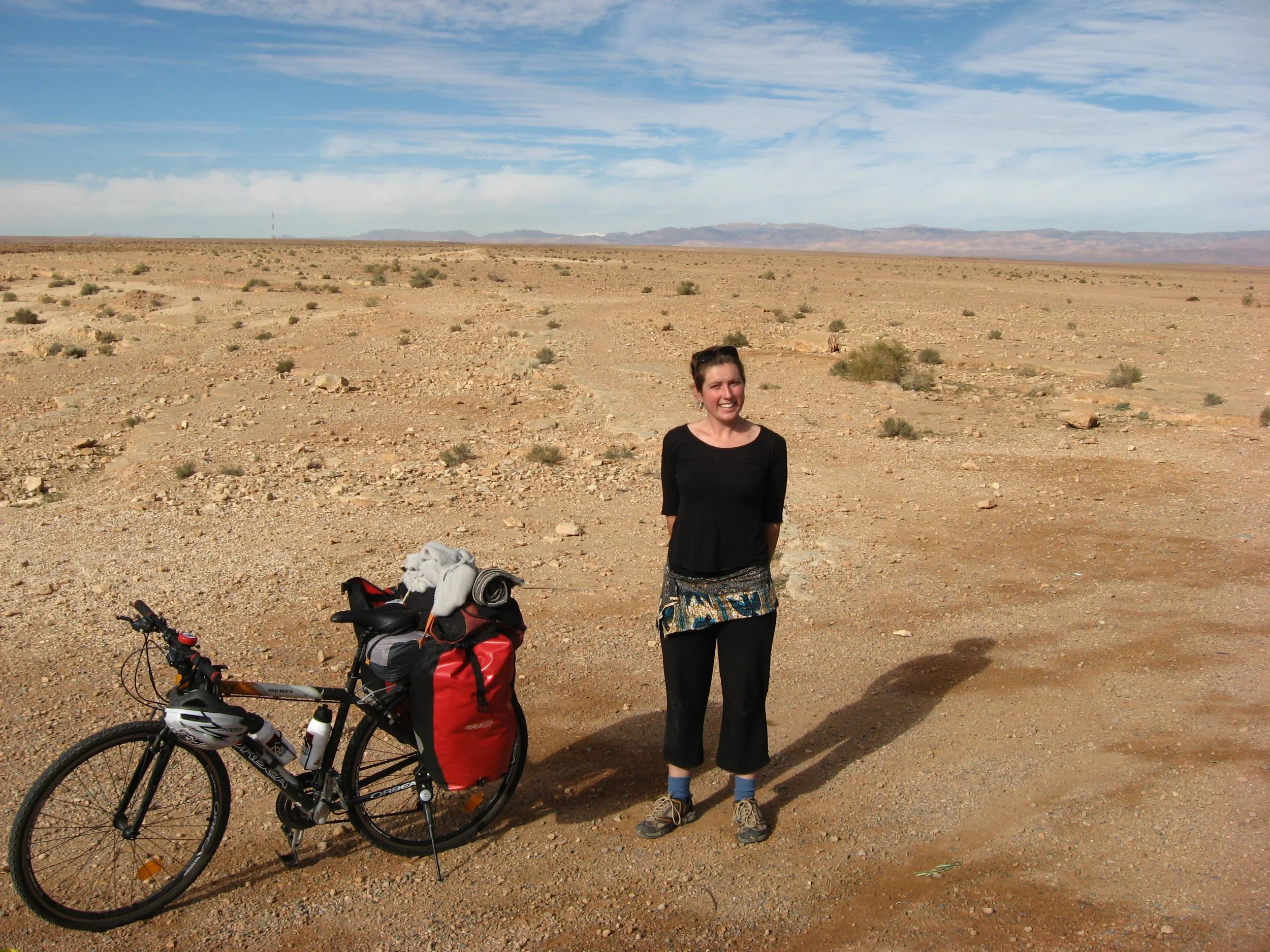 A woman (Sarah) standing in a barren desert landscape with a bicycle and loaded gear, smiling at the camera under a partly cloudy sky.