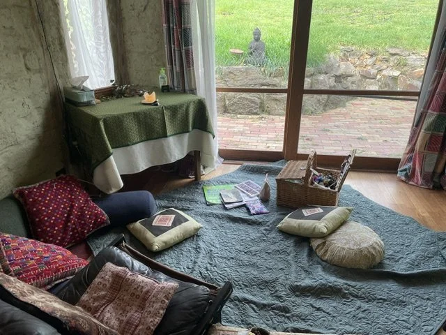 A cozy therapy room with cushions and blankets on the floor, a basket of art supplies and mindfulness cushion with a view of a garden with a Buddha statue outside through glass sliding doors.