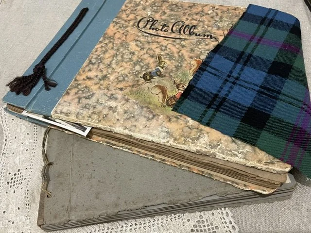 A stack of old photo albums and scrapbook albums, with a plaid cloth partially covering the top album, placed on a lace tablecloth.