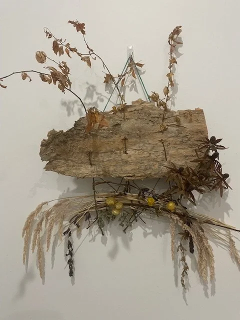 Mixed media wall art composed of a large piece of driftwood, dried leaves, and various dried plants arranged on a white wall.