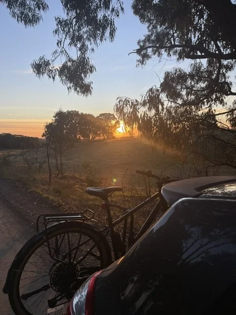 A sunset over a rural landscape with trees, a bicycle parked next to a car, and a dirt road.