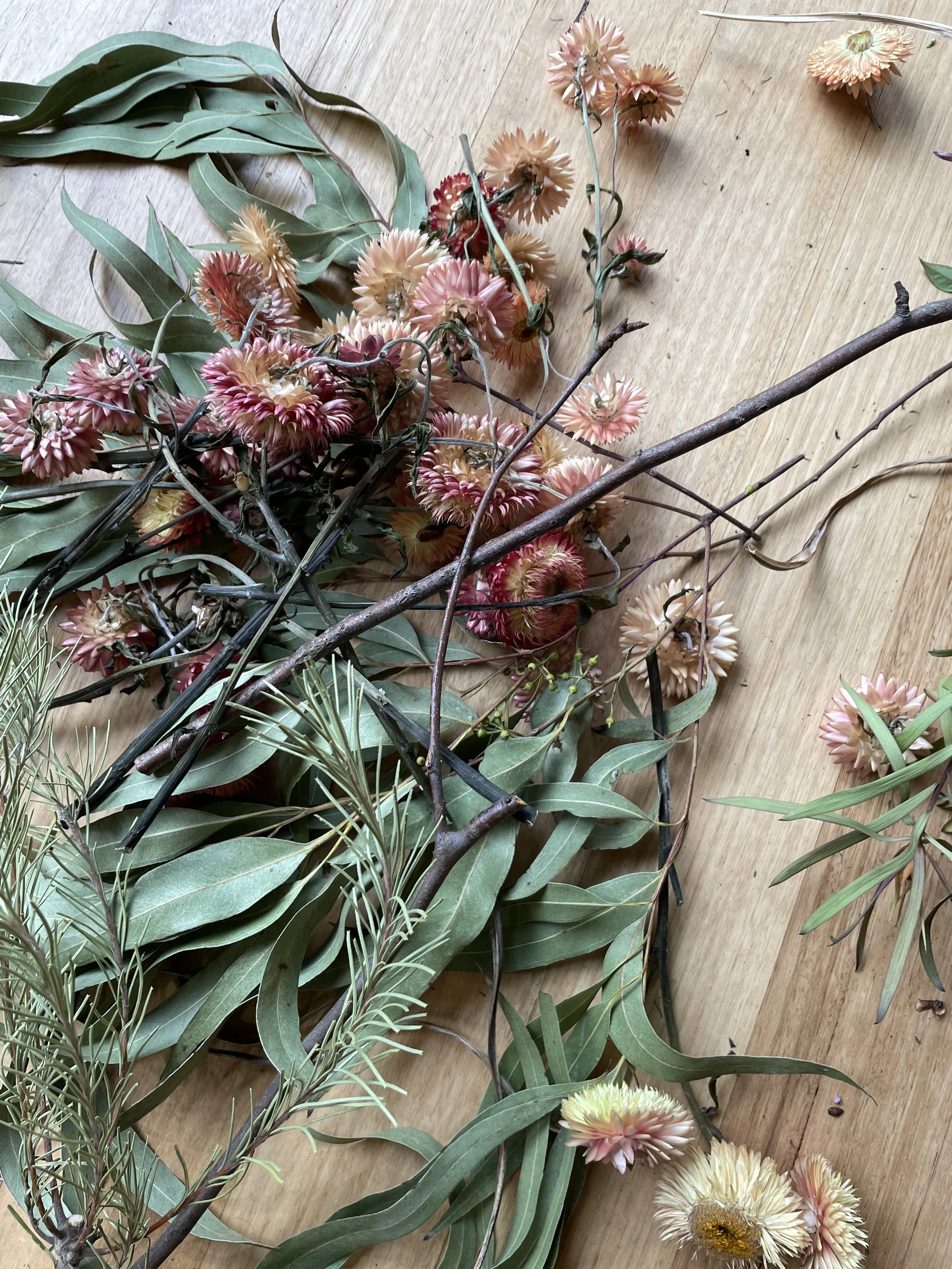 A variety of dried flowers and foliage, including pink and cream dahlias, long green leaves, and thin branches, all spread out on a wooden surface.