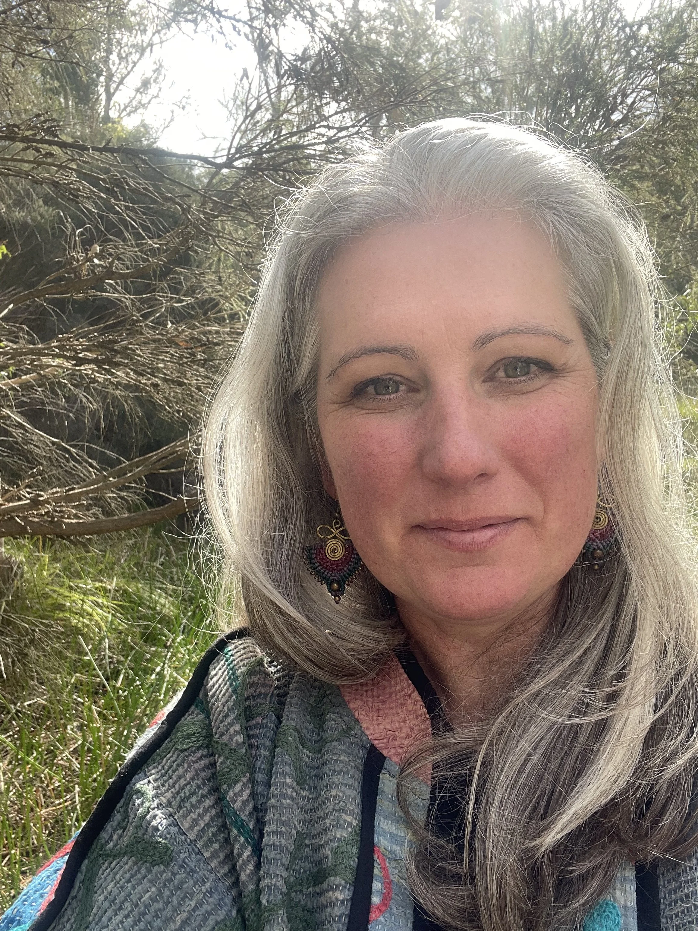 A woman, Sarah Brooke, with long gray hair and earrings standing outdoors in a natural setting. Warm facial expression, with sunlight and trees in the background.