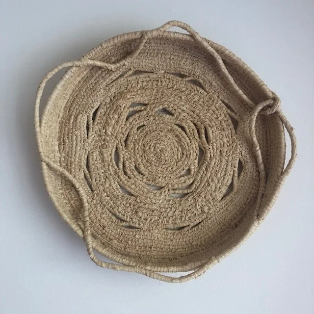 A round woven basket made of natural fibers with an open weave design, placed on a plain white surface.