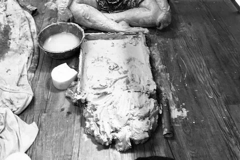 Person crossed legged on the floor with clay, wooden box, a bowl of water and a sponge.