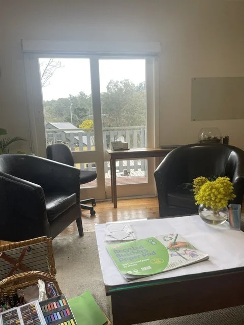 Light-filled therapy room with sliding glass door looking at tree-tops, two black armchairs, a small table with art supplies and fresh flowers.