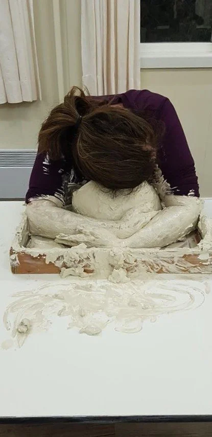 A person with brown hair leaning over a table, shaping a large lump of clay with their head and hands.