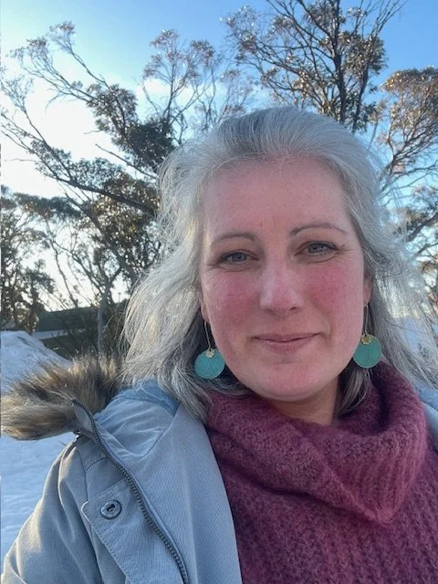 A woman (Sarah) with gray hair wearing turquoise earrings, a purple scarf, and a light blue jacket is outdoors in a snowy landscape with trees and a clear sky.