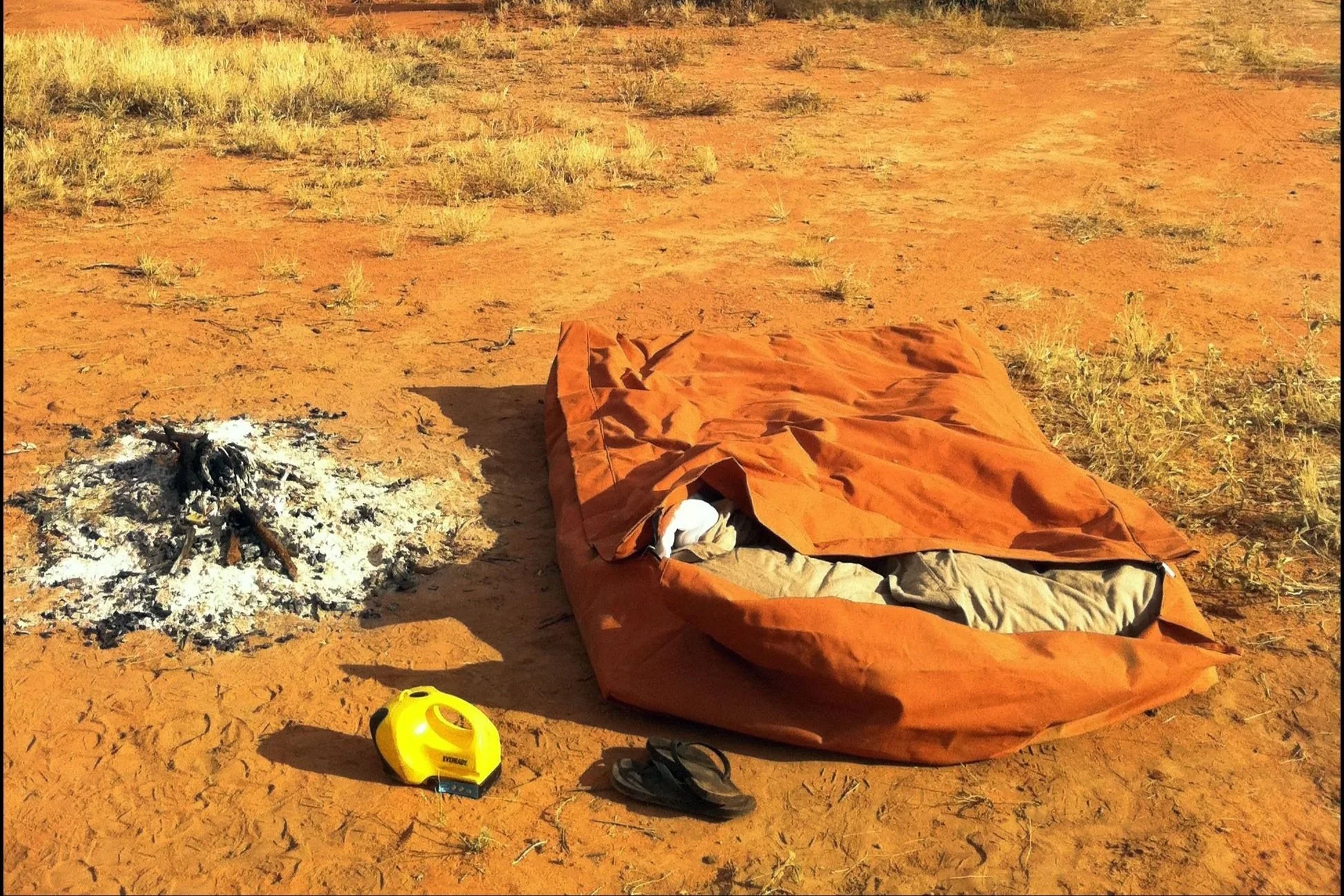 A s lying on the ground in a dry, open landscape with sparse vegetation. Nearby, there is a a yellow torch and some discarded items. There is also a small fire pit with ashes and charred wood.