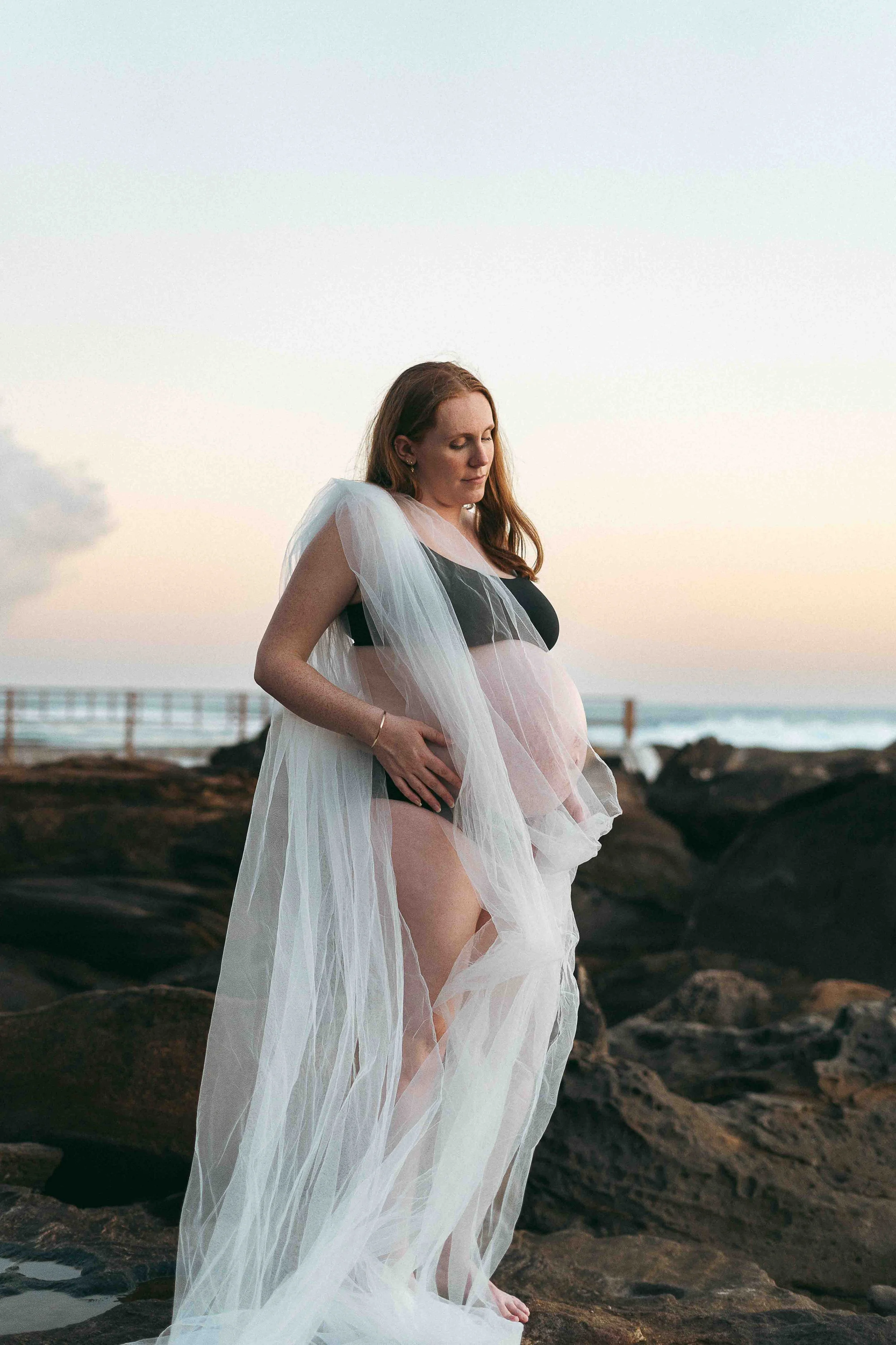 A woman in a flowing dress standing on rocks near the water during sunset with her eyes closed.