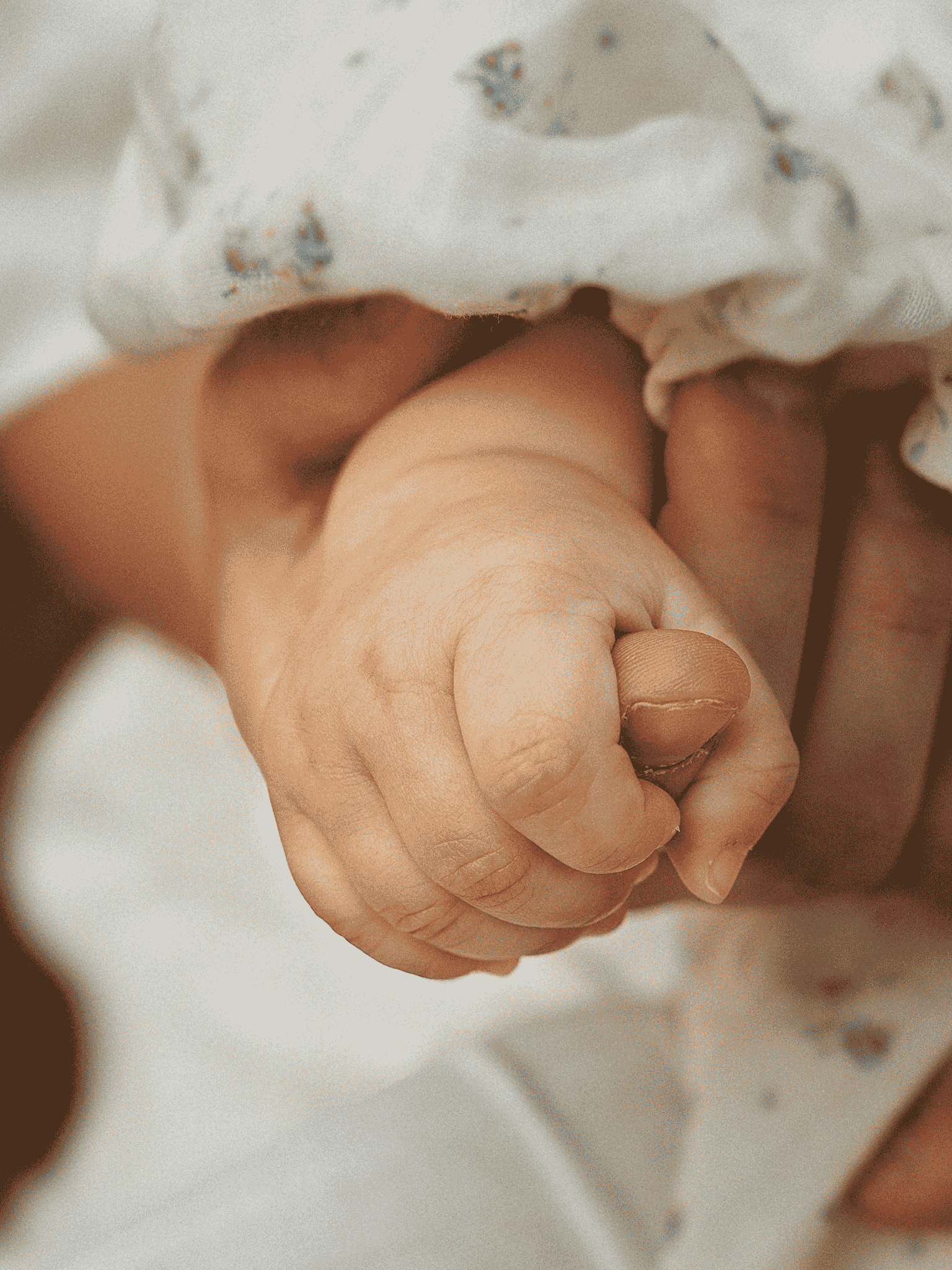 Close-up of a person holding a baby's hand, with a blanket covering part of the hand. The baby is wrapped in a light-colored blanket with small blue and gray patterns.