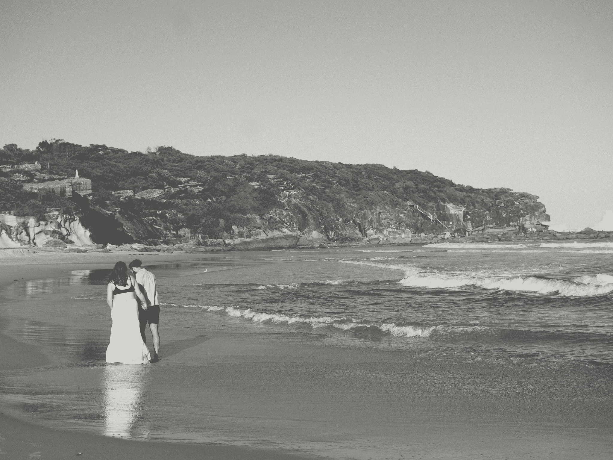 A black and white photograph of a beach scene, showing a couple walking along the shoreline holding hands. In the background, there are rocky cliffs with some vegetation and a staircase leading up the cliffside. The sky appears clear, and the ocean waves are gently crashing onto the shore.