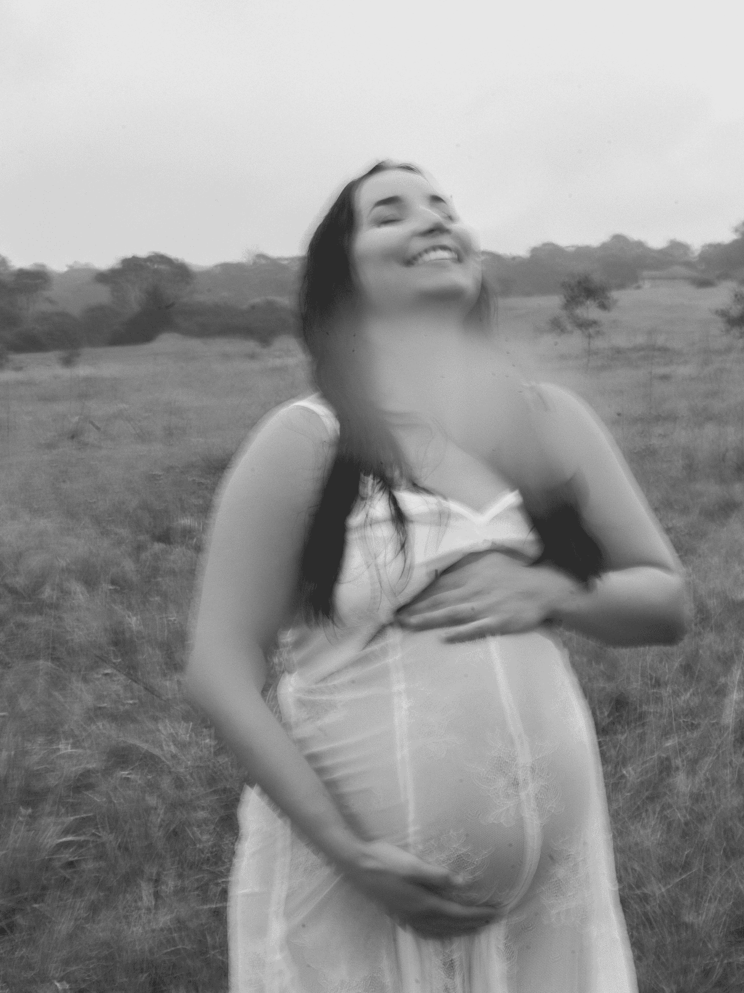 A young woman standing outdoors in a field with trees in the background, smiling with her eyes closed, wearing a dress with sleeveless top and her hair down.