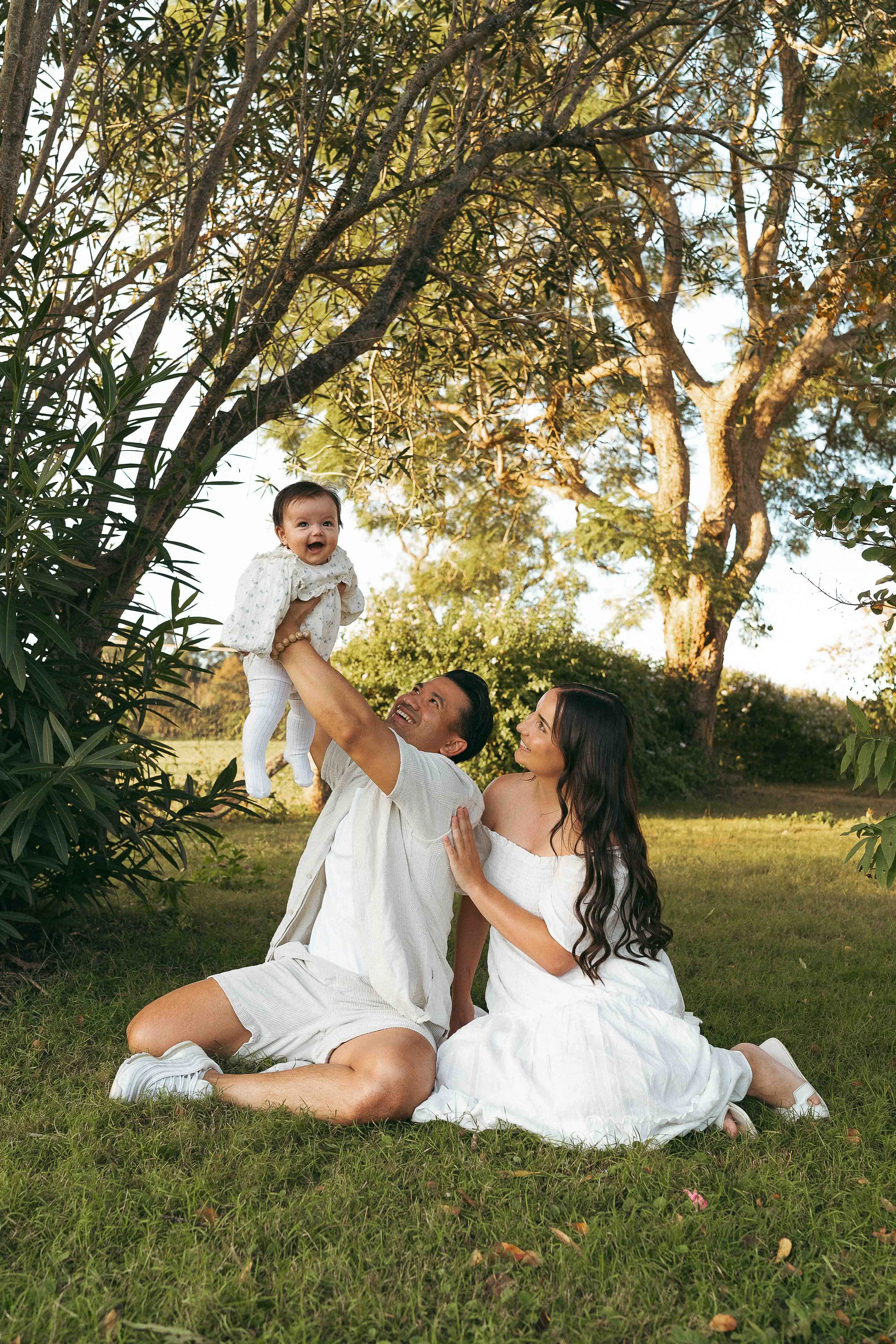 Family of three outdoors in a grassy park, with a large tree and clear sky in the background. The man is sitting on the grass holding a young girl, and a woman is sitting beside him. All are smiling and enjoying the moment.