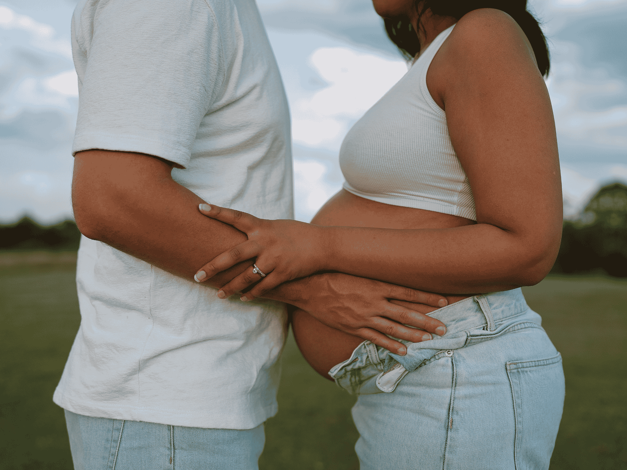 A pregnant woman in a white tank top and denim shorts touching her belly, standing close to a man in a white t-shirt, with her hand resting on their shared belly area. They are outdoors in a grassy area under a cloudy sky.