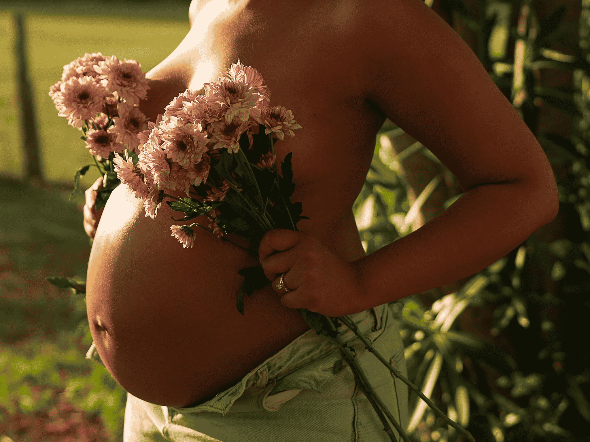 A person from the neck down, holding a bouquet of pink flowers close to their chest, outdoors with sunlight.